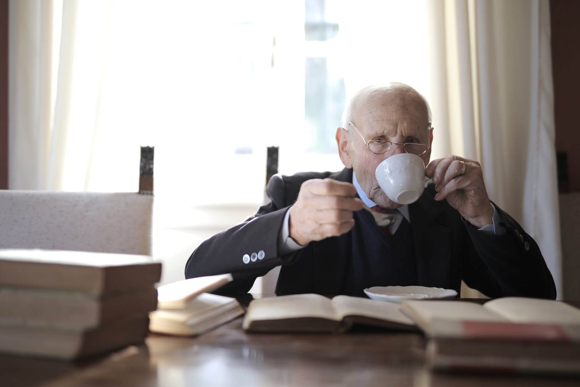 senior man drinking hot beverage while sitting at table with books