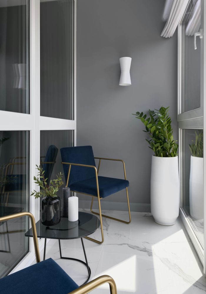 Modern balcony design featuring two blue armchairs with gold frames, a small black side table, and a tall white planter with a green plant.