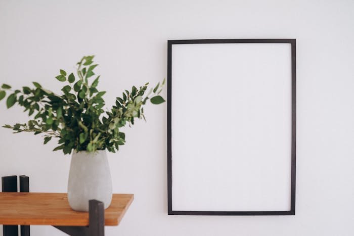 An empty black frame beside a green plant in a concrete vase on a wooden shelf.