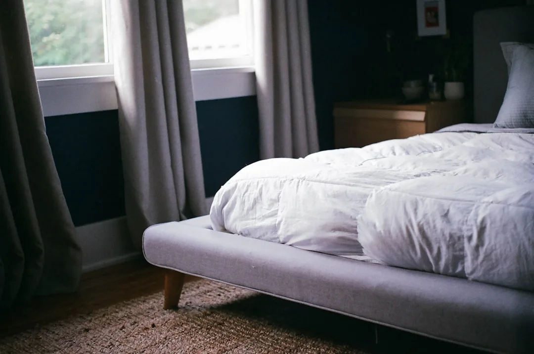 Low-profile platform bed with white linen duvet near window in dark-walled bedroom with wood nightstand