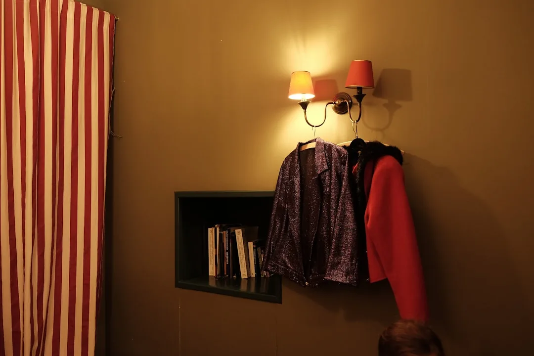 Wall-mounted double sconce with warm light above built-in shelf with books and hanging jackets in small room