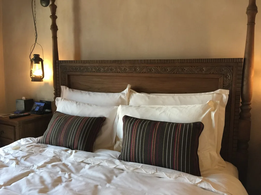 Layered white hotel-style bedding with striped accent pillows on carved wooden four-poster bed with lantern lighting
