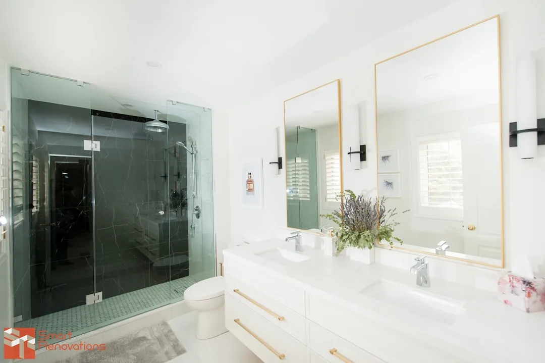 Walk-in glass shower with dark marble tile walls in modern white bathroom with double vanity and gold mirrors