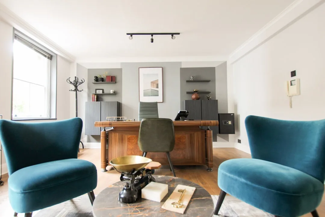 Stylish home office with wooden executive desk, teal velvet chairs, and gray accent wall in a compact room