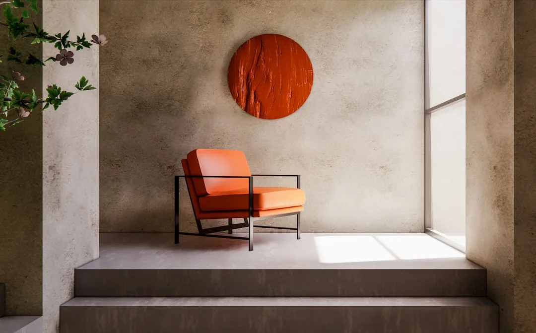 Large circular orange wall sculpture above a modern metal-frame chair in a minimalist concrete interior space