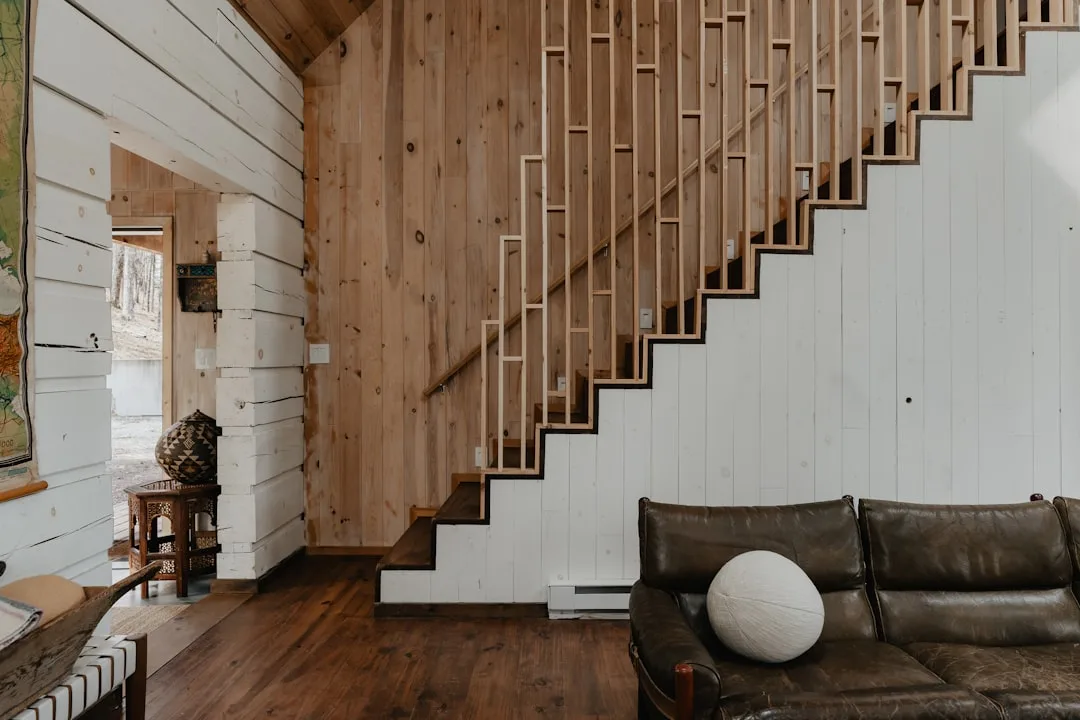 Open-plan living room with wooden staircase showing triangular under-stair space beside leather sofa in rustic cabin