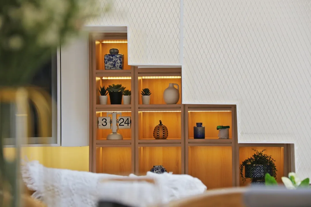 Built-in wooden shelving unit with LED lighting displaying decorative vases, plants, and clock in a modern interior