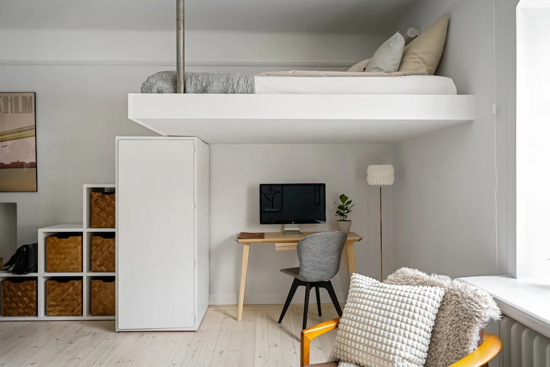 Renter-friendly loft bedroom with freestanding wooden desk, gray chair, and white shelving unit in small studio apartmen