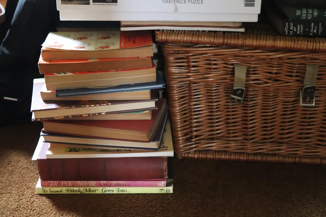 Stack of hardcover books piled on floor beside wicker storage basket with buckle straps