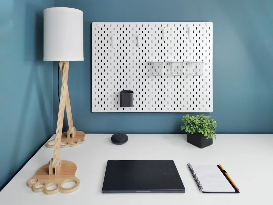 White pegboard wall organizer with storage bins above a minimalist desk with lamp, notebook, and laptop