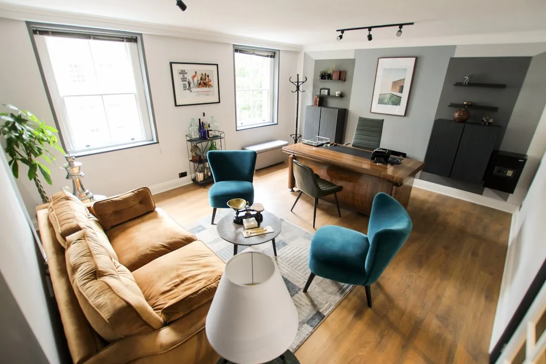 Small home office with wooden desk, teal velvet chairs, brown sofa, and track lighting on hardwood floors