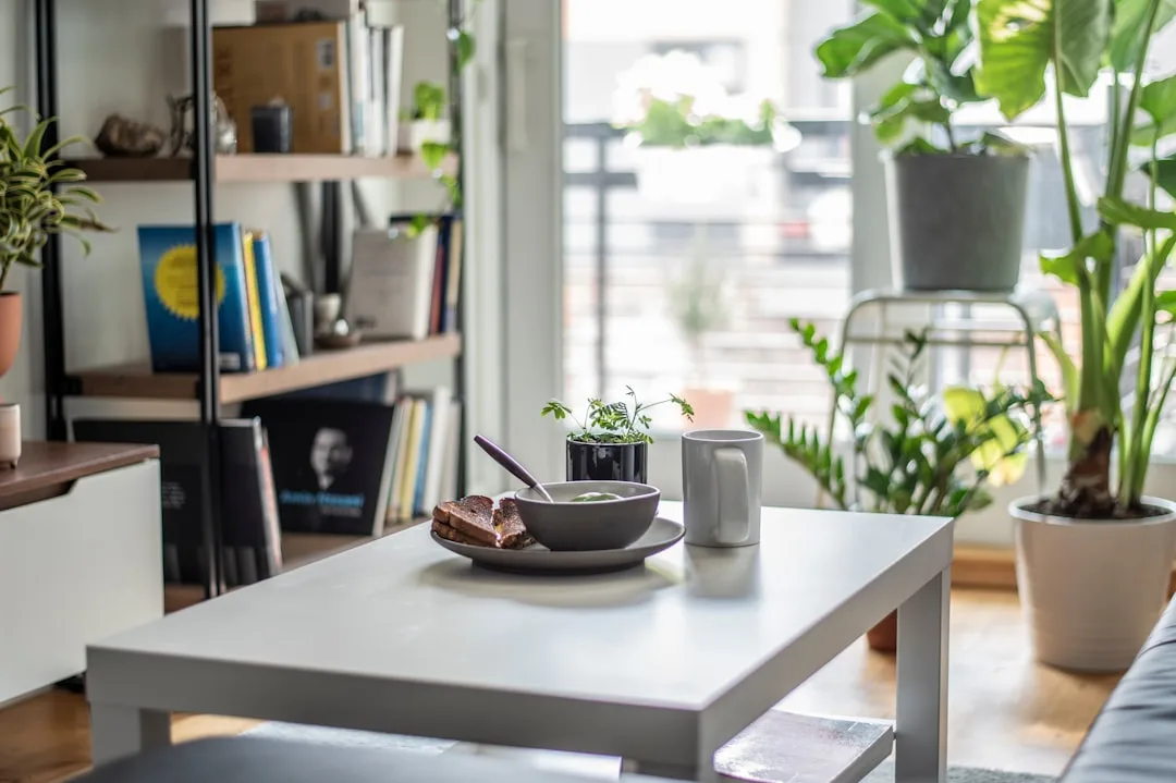 Small white coffee table with bowl and mug in a cozy living room surrounded by indoor plants and bookshelves