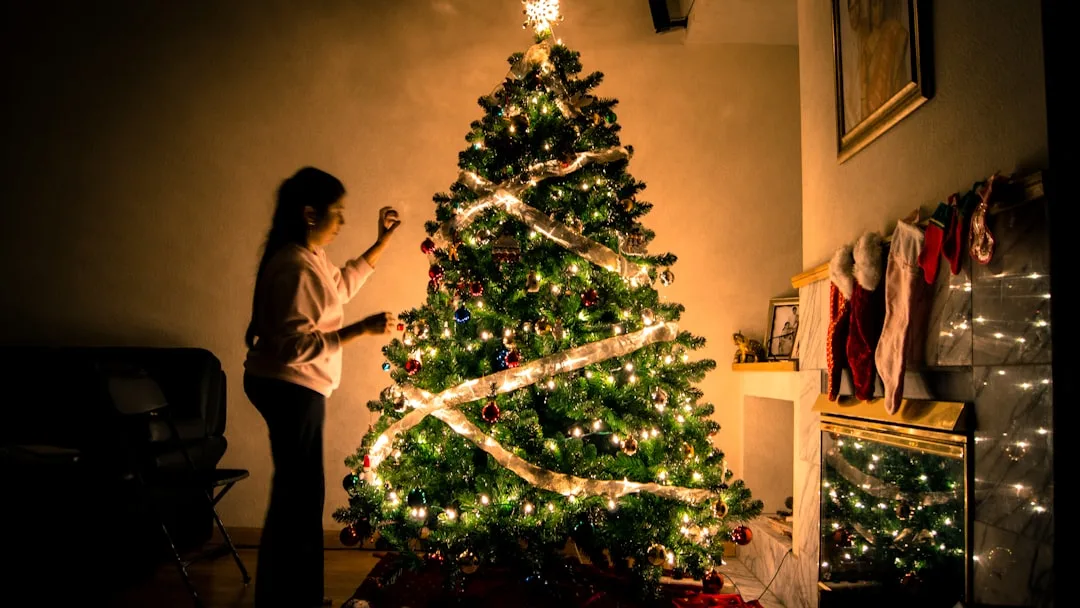 Woman decorating a large lit Christmas tree with ornaments and ribbon in a cozy living room with stockings by fireplace