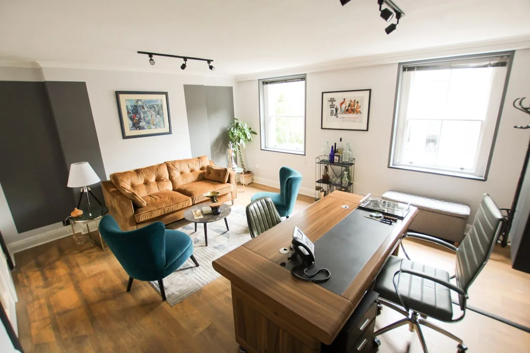 Dual-function office and lounge space with wooden desk, teal accent chairs, velvet sofa, and track lighting