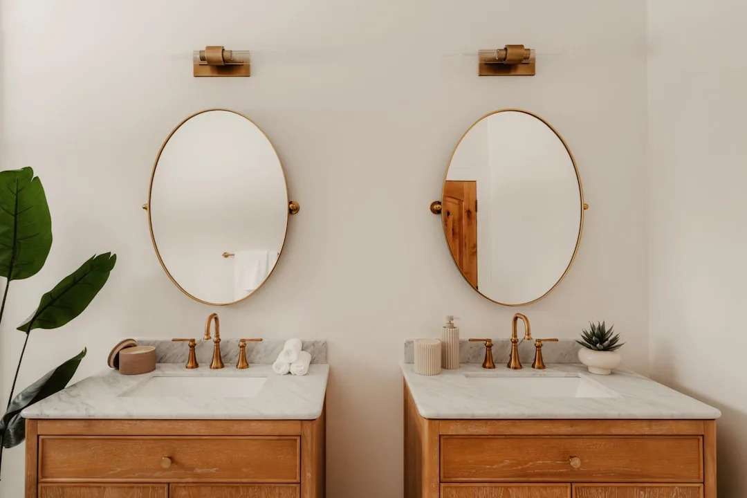 Double bathroom vanity with gold oval mirrors, marble countertops, and brass faucets mounted at standard height on white