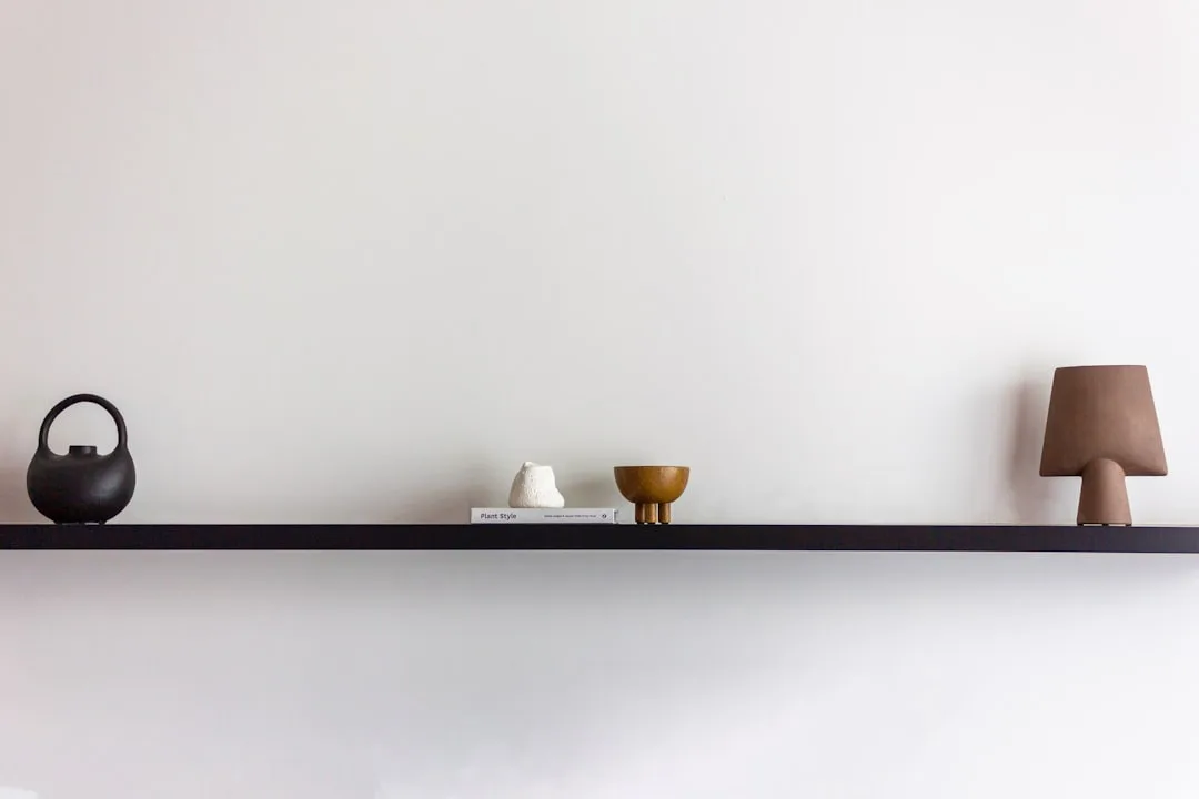 Minimalist floating shelf with black ceramic vase, white sculpture, wooden bowl and small lamp against white wall