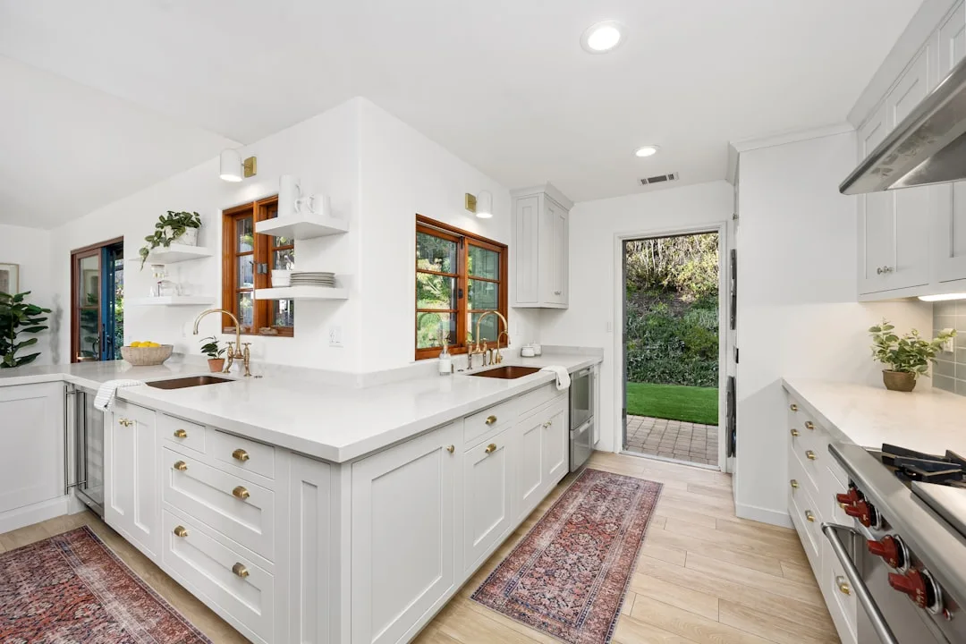 White kitchen cabinets with gold hardware, quartz countertops, and warm wood-framed windows in a bright modern kitchen