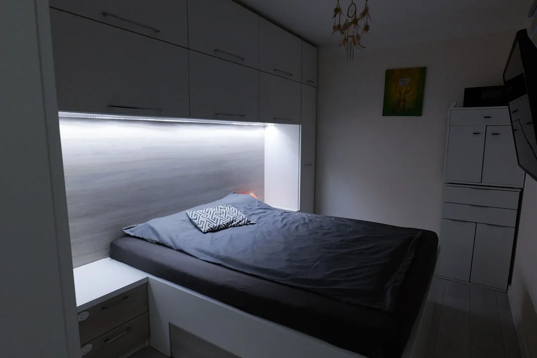 Small bedroom with LED strip lighting under overhead cabinets illuminating a built-in bed in a compact apartment