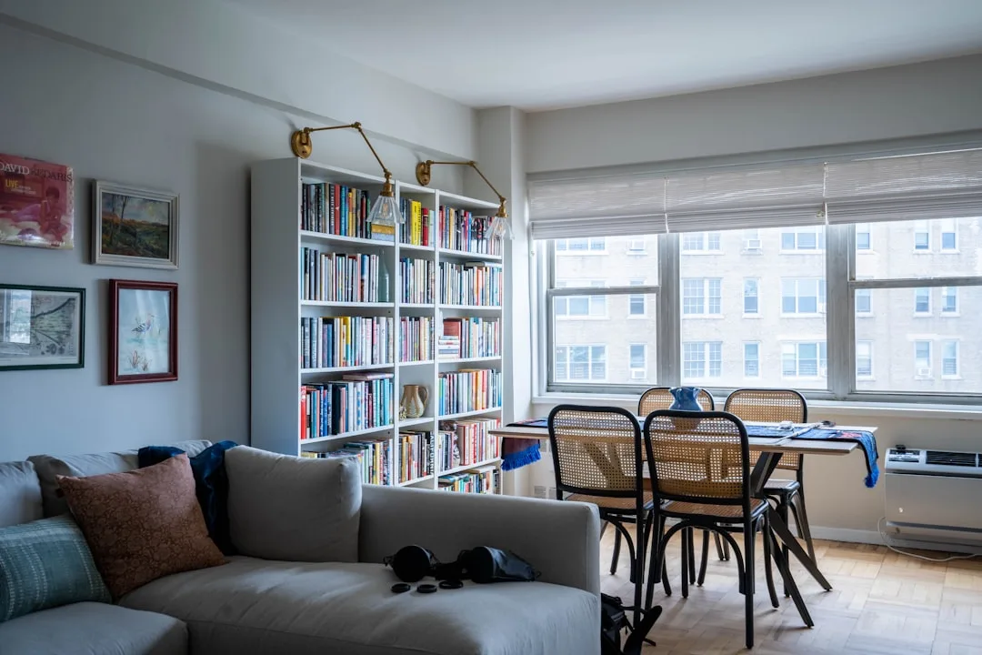 Budget rental apartment living room with white bookshelf, gray sofa, dining table and large windows in NYC apartment