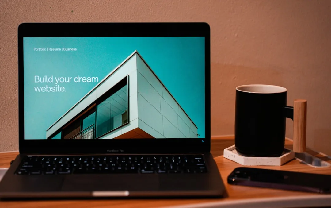 MacBook Pro displaying a website builder homepage on a wooden desk beside a black coffee mug and smartphone