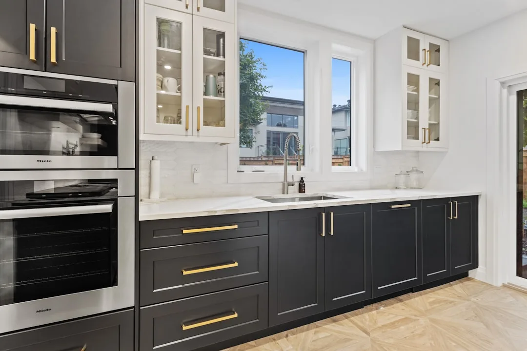 Dark gray shaker kitchen cabinets with gold hardware and white uppers flanking Miele double wall ovens and marble counte