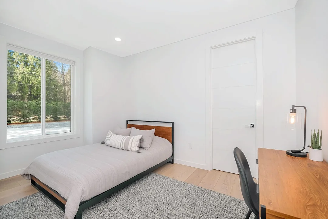 Small bedroom with window and door on same wall, single bed, wood desk, light hardwood floors and white walls