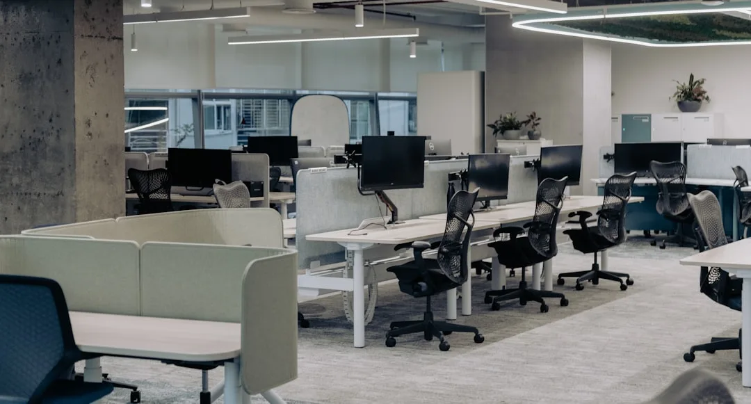 Modern open-plan office with white sit-stand desks, black ergonomic chairs, and monitor arms in a professional workspace