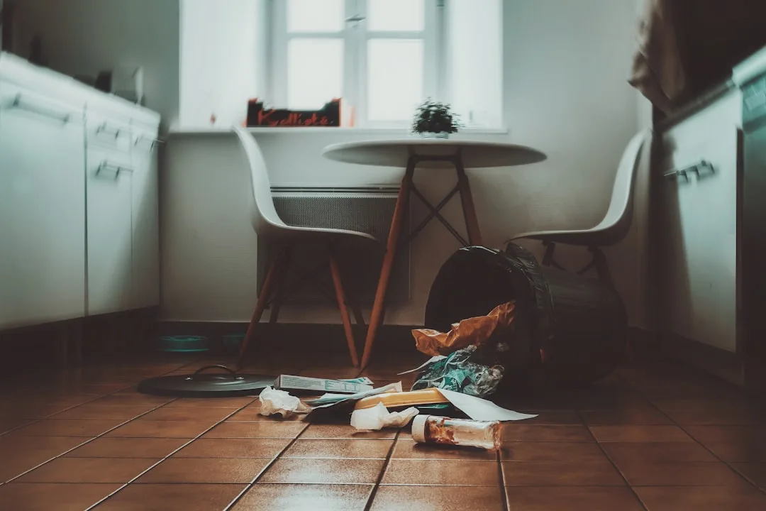Overturned trash can with spilled garbage on terracotta kitchen floor tiles near white Eames chairs and table
