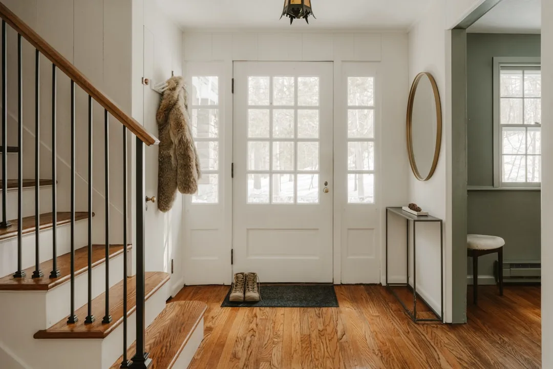 Clean farmhouse entryway with white front door, wood floors, staircase, round mirror, and fur coat hanging on hook