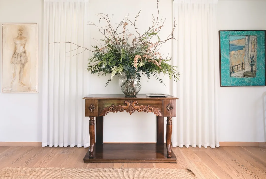 Ornate carved dark wood console table with large floral arrangement, flanked by artwork on white walls with linen curtai
