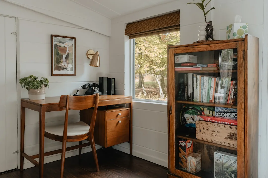 Wooden bookshelf cabinet with board games and books beside a mid-century desk in a cozy cabin workspace corner