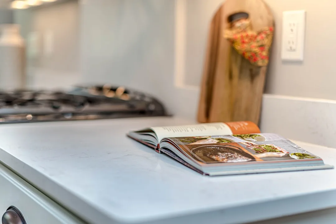 Open cookbook on white marble kitchen island countertop with gas range and wooden cutting board in background