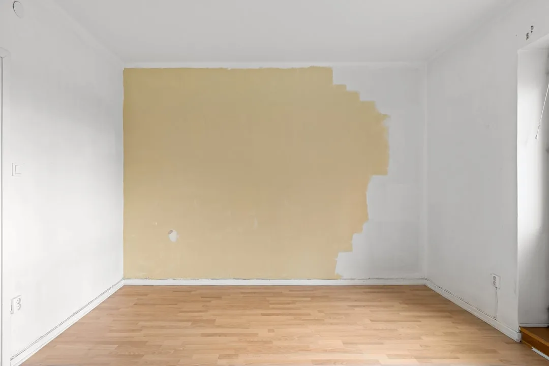 Empty room with partially stripped beige wall, white baseboards, and light wood laminate flooring during home renovation
