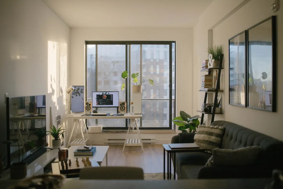 Work from home setup in rental apartment with white desk, monitor, speakers, sofa, and plants near large window