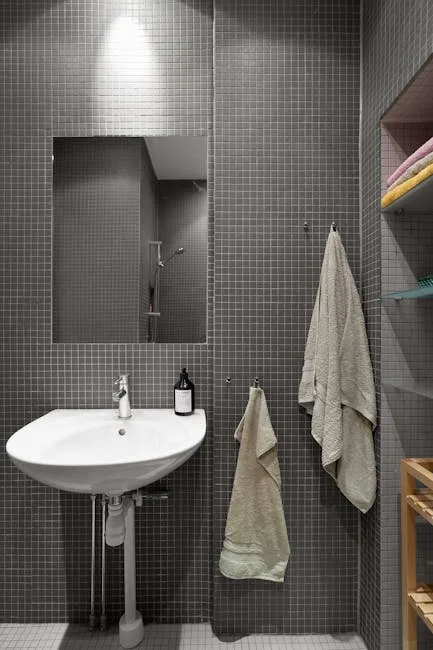 Small bathroom with gray mosaic tiles, pedestal sink, wall mirror, hanging towels, and open shelving for storage