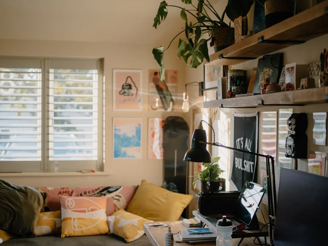 Cluttered living room workspace with shelves, plants, posters, pillows and desk items in a cozy eclectic home