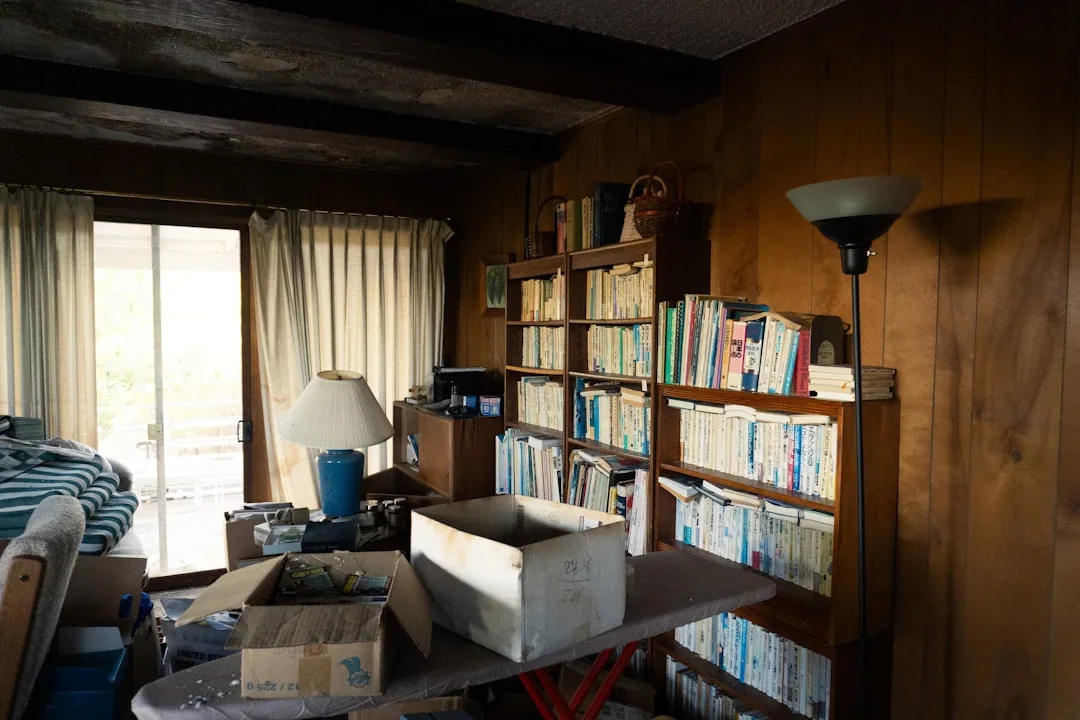 Cluttered wooden-paneled room with overflowing bookshelves, cardboard boxes, and mismatched furniture creating a disorga