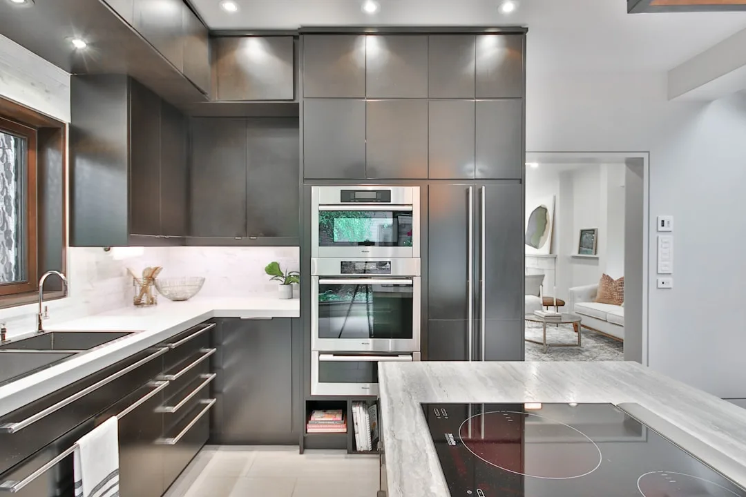 Modern kitchen remodel with dark gray custom cabinetry, stainless steel built-in ovens, marble countertops, and inductio