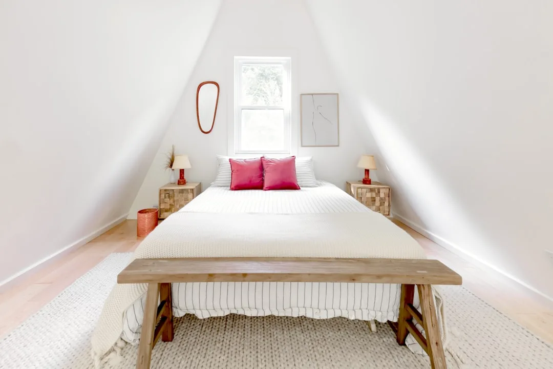 Converted attic bedroom with sloped white ceilings, wood floor, bed with pink pillows and wooden bench at foot