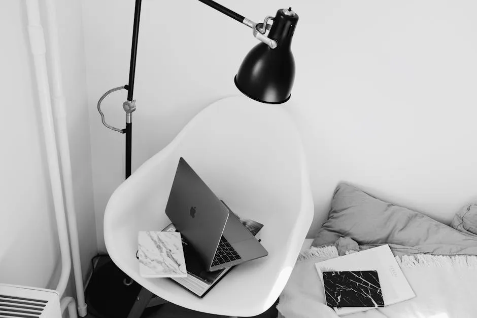 MacBook laptop on modern white chair beside bed with black floor lamp in minimalist bedroom corner home office setup