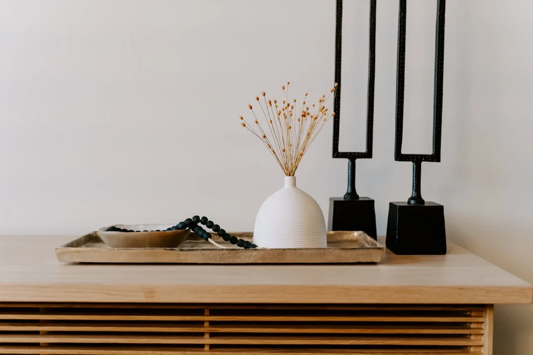 Wooden tray with white ceramic vase, dried flowers, beaded bracelet, and black decorative candleholders on a slatted con