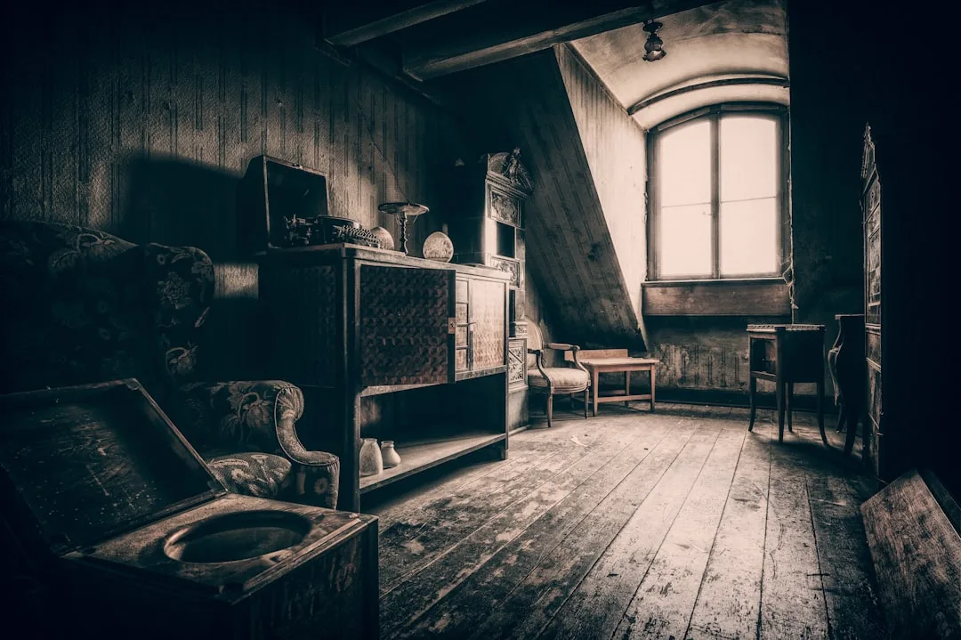Dark vintage attic space with sloped ceiling, wooden floors, antique furniture and arched window showing conversion pote
