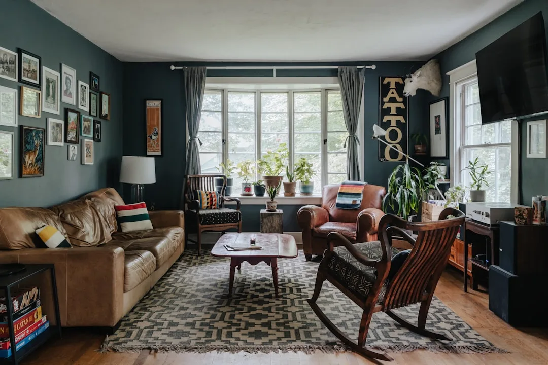 Eclectic living room with dark teal walls, brown leather sofa, rocking chair, and mixed vintage furniture on patterned r