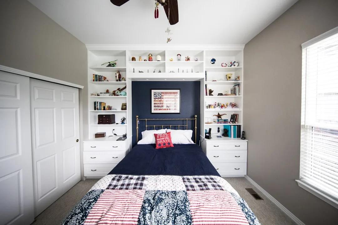 Small bedroom with built-in white shelving units, dual dresser drawers, and navy blue bedding maximizing storage space