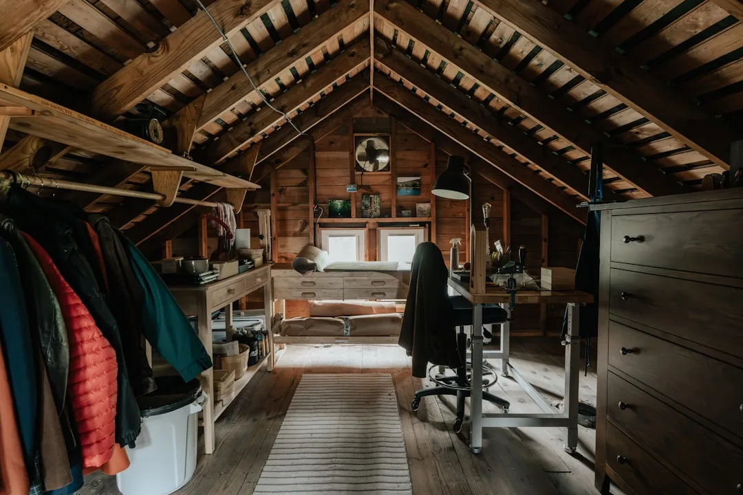 Converted attic room with wood beams, desk workspace, clothing rack, and bed showing finished living space conversion