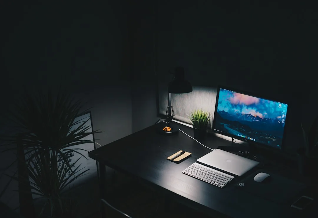 Minimalist work from home desk setup with monitor, MacBook, keyboard, mouse, and desk lamp in dark room