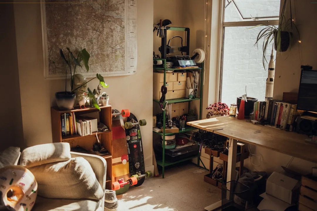 Cluttered home workspace with shelves, skateboards, plants, books, and a 3D printer near a bright window
