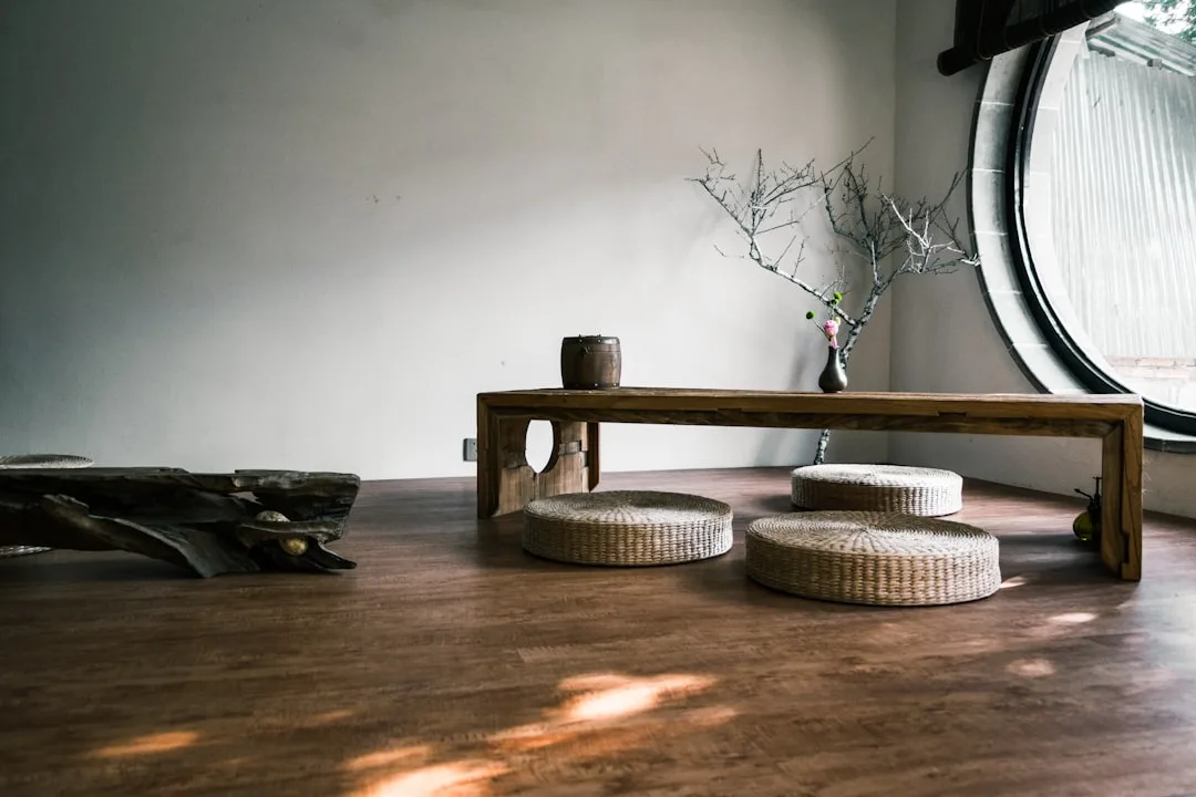 Japandi-style low wooden bench with woven floor cushions, branch arrangement in vase, and round window in minimalist roo