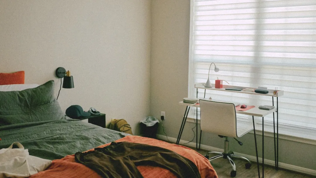 Minimalist dorm room with green bedding, white desk, chair, and window blinds showing a simple student bedroom aesthetic