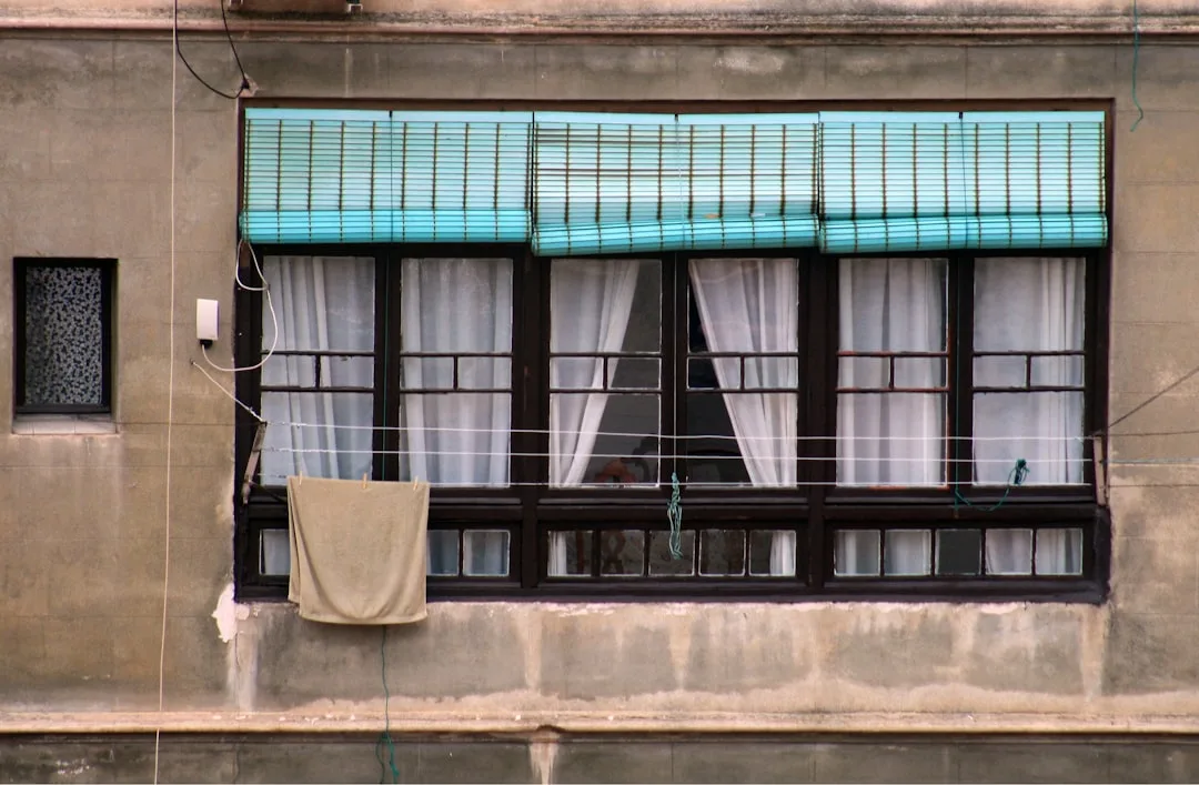 Teal bamboo roman shade over builder-grade flat windows with white sheer curtains on aging concrete building facade